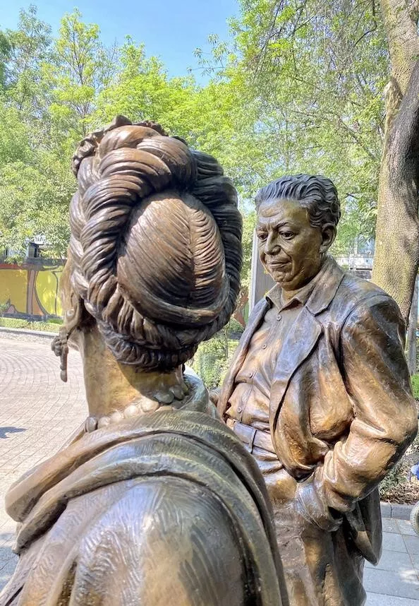 Two statues are captured in a tree-filled plaza. One statute is a man white short wavy hair in a blazer looking down. The woman statute’s picture is taken from behind. Her braided hair is wrapped around her head, she is wearing earrings, a large beaded necklace, and a shawl on her shoulders.