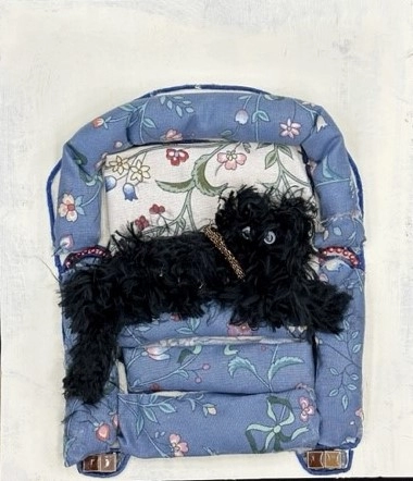 This whimsical tactile collage features a furry black dog made of yarn seated comfortably on a primarily light blue upholstered chair with a floral pattern. The feet of the chair are made of two brown ceramic tiles each. The background is white and it in a black vertical rectangular frame.