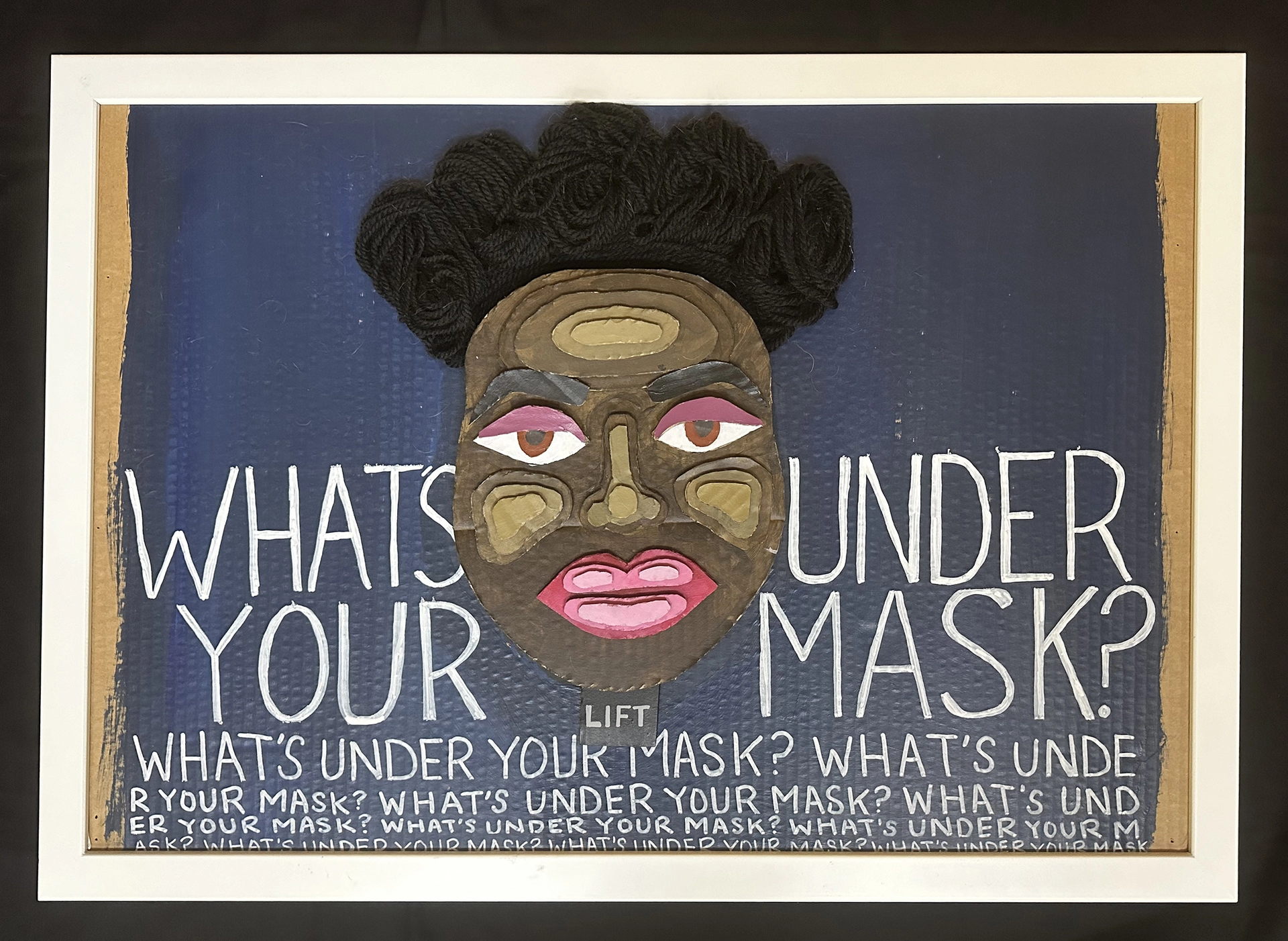 The cardboard texture shows through the blue background. In white text, the question "What is under your mask" is repeated. The center features a face made of layered cardboard, with a medium brown tone, pink lips, pink eyelids, and crowned with black yarn hair.