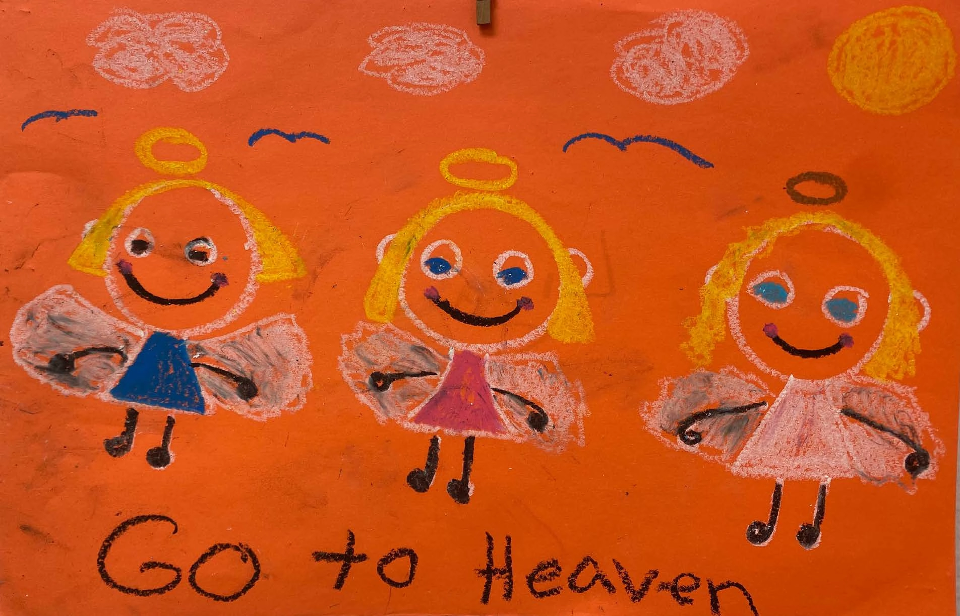 On orange paper, a simple drawing features three smiling blonde angels with wings and halos floating among blue birds, white clouds, and a round sun. The text at the bottom reads "Go to Heaven."