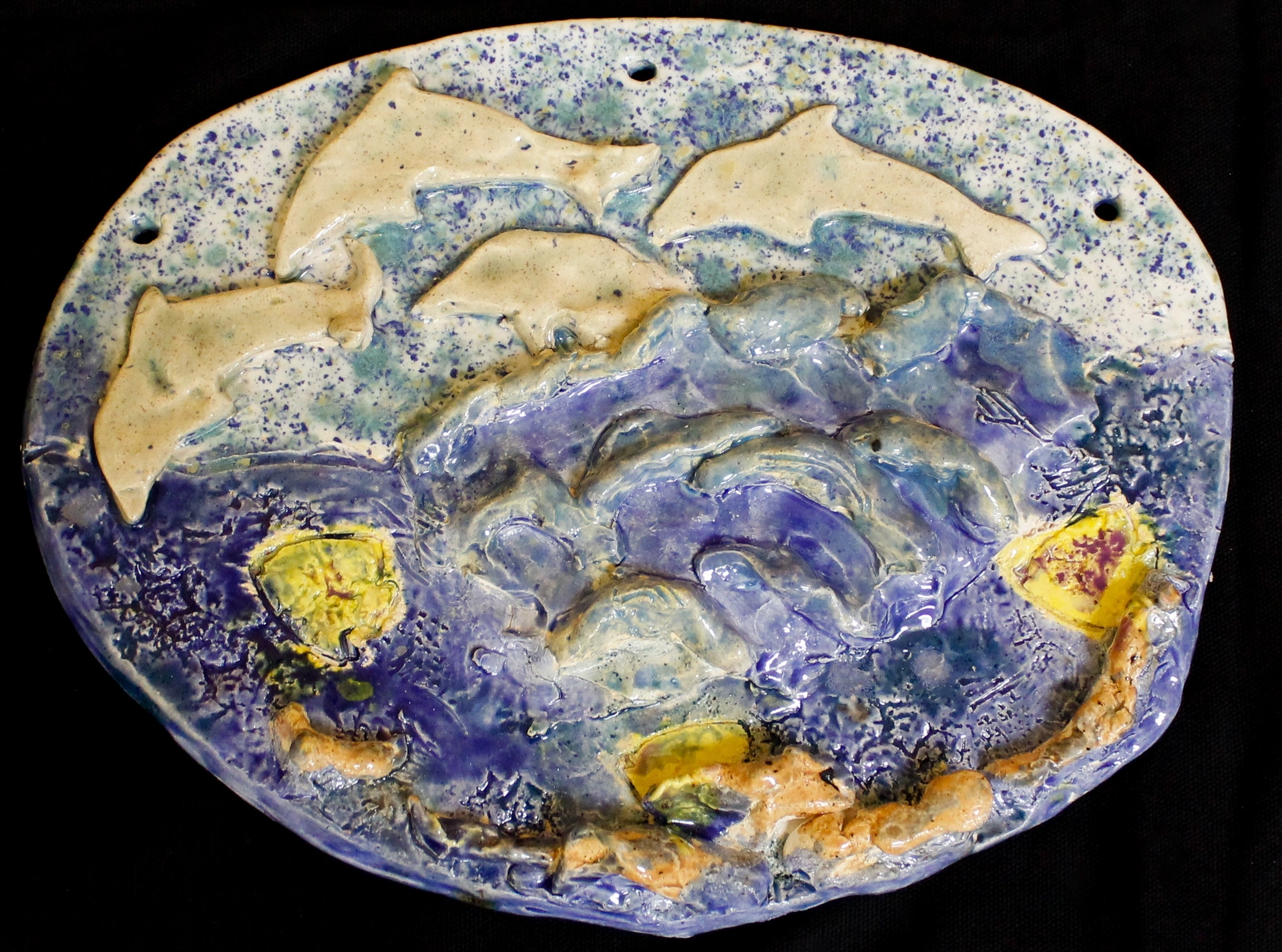 A photograph of a ceramic plaque in a horizontal oval depicts a pod of yellow dolphins breaching the blue sea below.