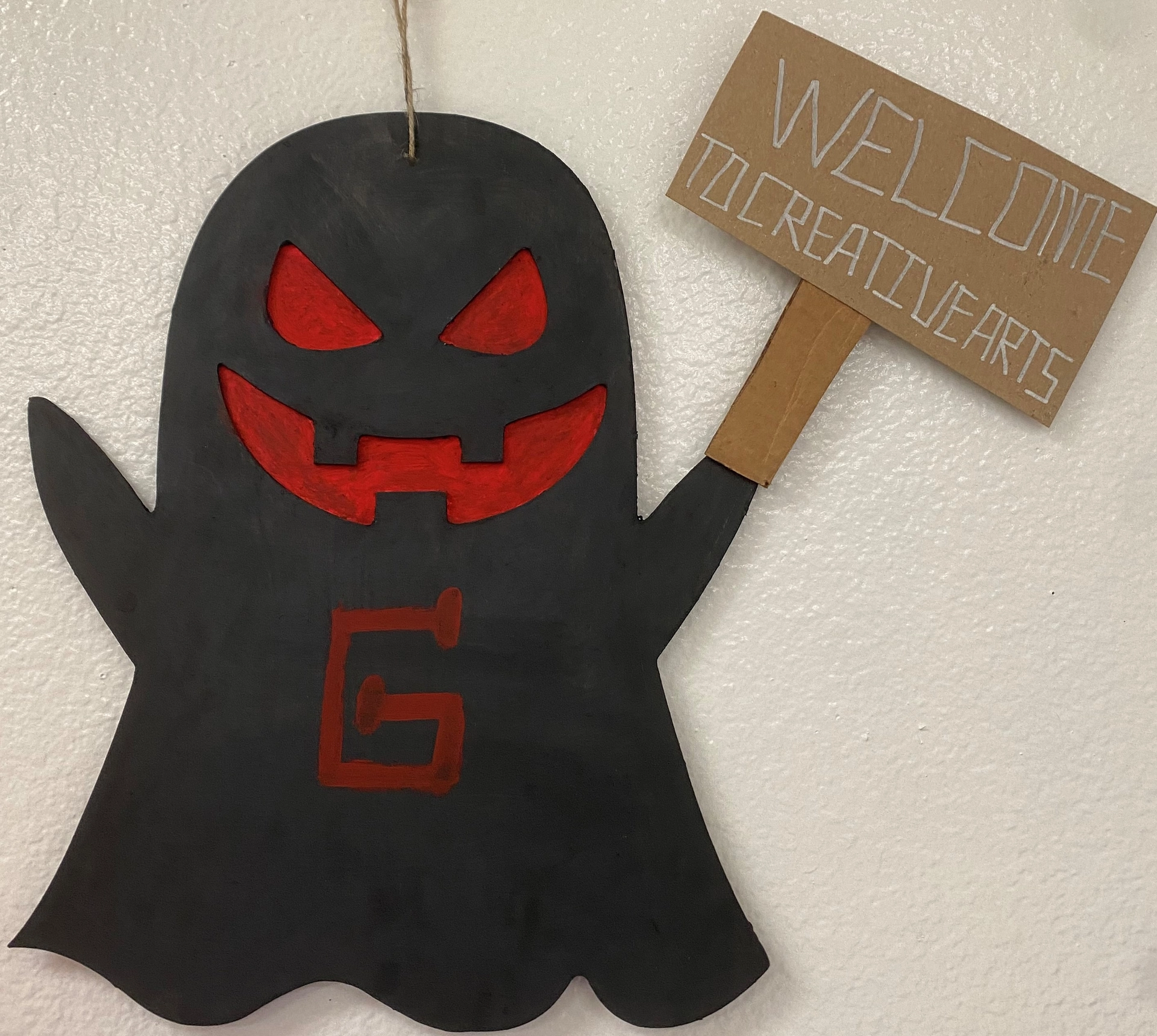A photograph of a black ghost-shaped cutout with red eyes and mouth, in cut-out relief, features a red letter "G" in the center and holds a sign that reads "Welcome to Creative Arts."