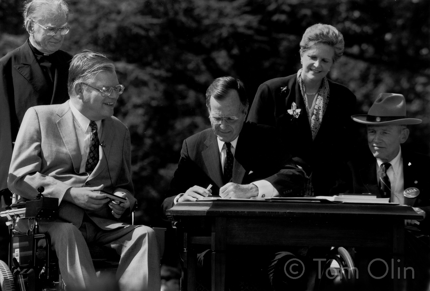 Black and white photo of President George H.W. Bush seated at a wooden desk outside, signing the Americans with Disabilities Act (ADA) into law on July 26, 1990. The image is credited to photographer Tom Olin, whose watermark appears in the bottom right corner.
