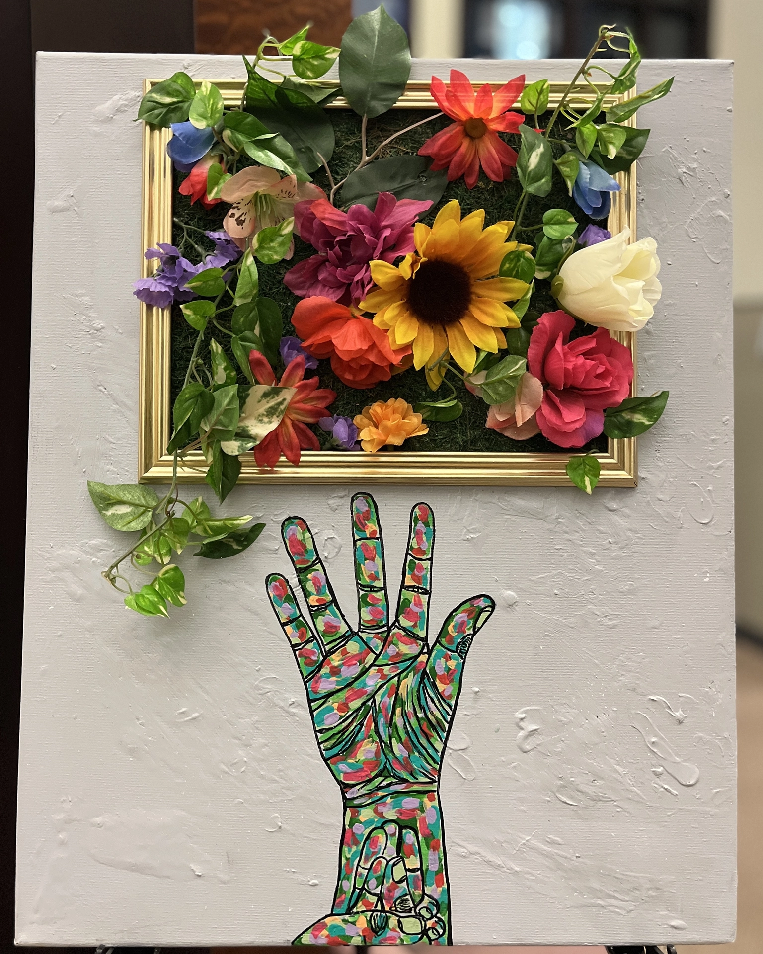 A textured gray canvas features a gold frame filled with colorful artificial flowers on a background of green moss. Below the frame, a raised hand is illustrated in an American Sign Language (ASL) handshape that spells “GROWTH.” The hand is painted with vibrant brushstrokes in red, green, yellow, and purple.