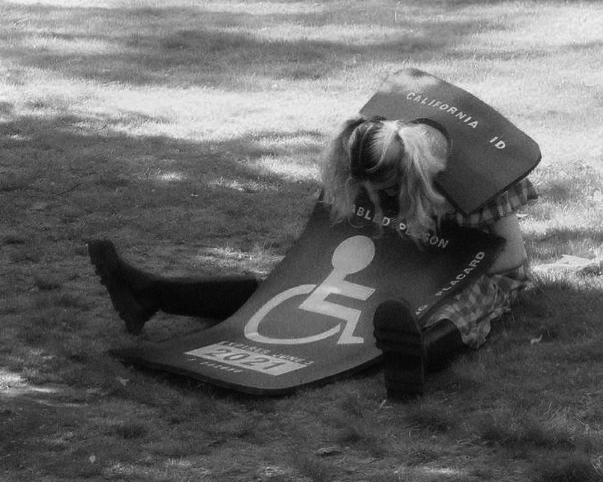 A girl is sitting on the grass looking down, being weighed down by a sculpture of a life-size disability parking pass around her neck.