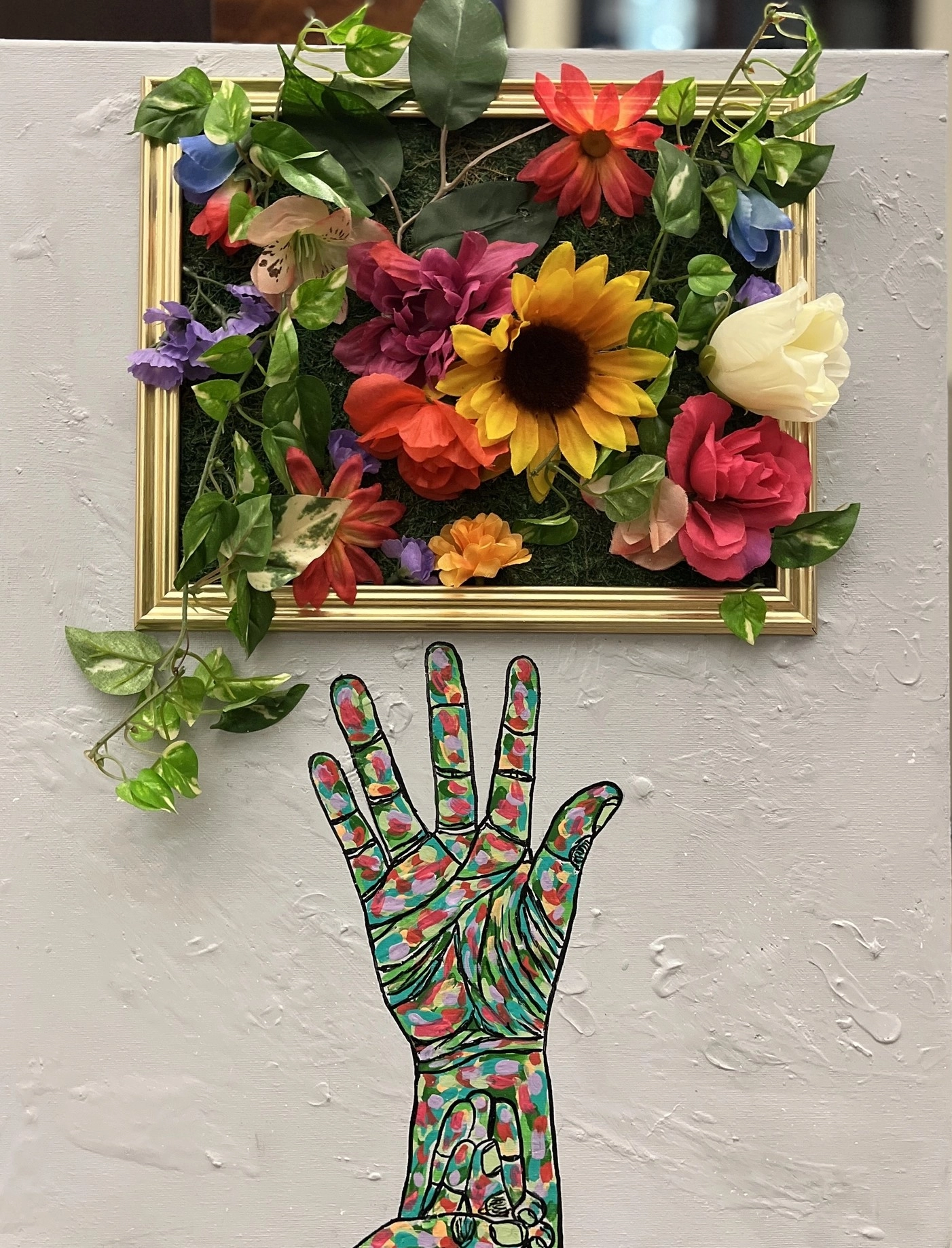 A gray textured canvas holds a gold frame filled with colorful artificial flowers and green moss. Below the frame, a hand is illustrated in American Sign Language spelling "GROWTH," painted with vibrant strokes of red, green, yellow, and purple.