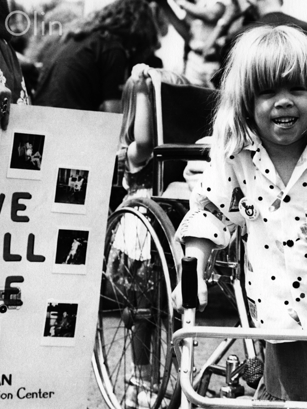 A young child with blonde hair and a walker smiles at the camera during a protest. Next to her, someone holds a poster that says “We Will Ride” from the Lowman Special Education Center, demanding accessible transportation.