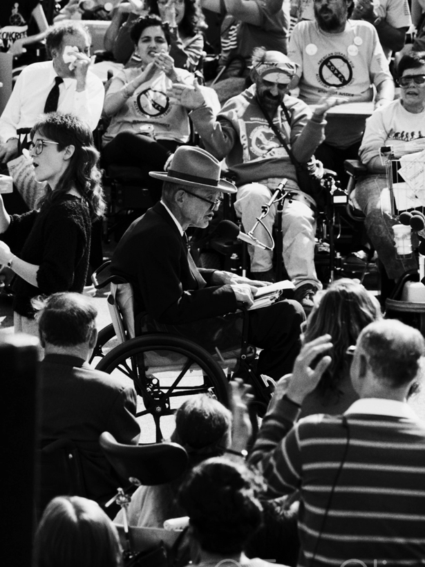 Justin Dart Jr., wearing his signature cowboy hat and seated in a wheelchair, speaks into a microphone at a rally. Behind him, a crowd of disabled activists cheer and applaud, many wearing ADAPT T-shirts.