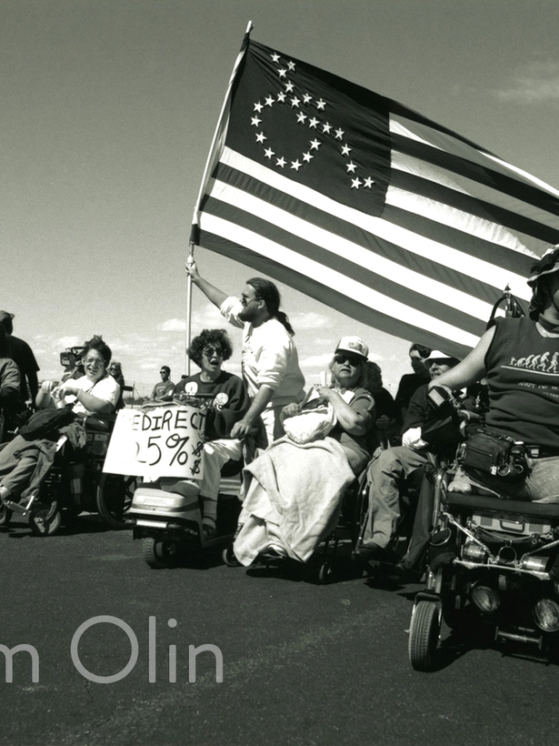 A large group of disability rights activists, most using wheelchairs, line up across a city street holding signs, including one that reads “Redirect 25%.” An adapted American flag with the disability access symbol in the stars flies overhead.
