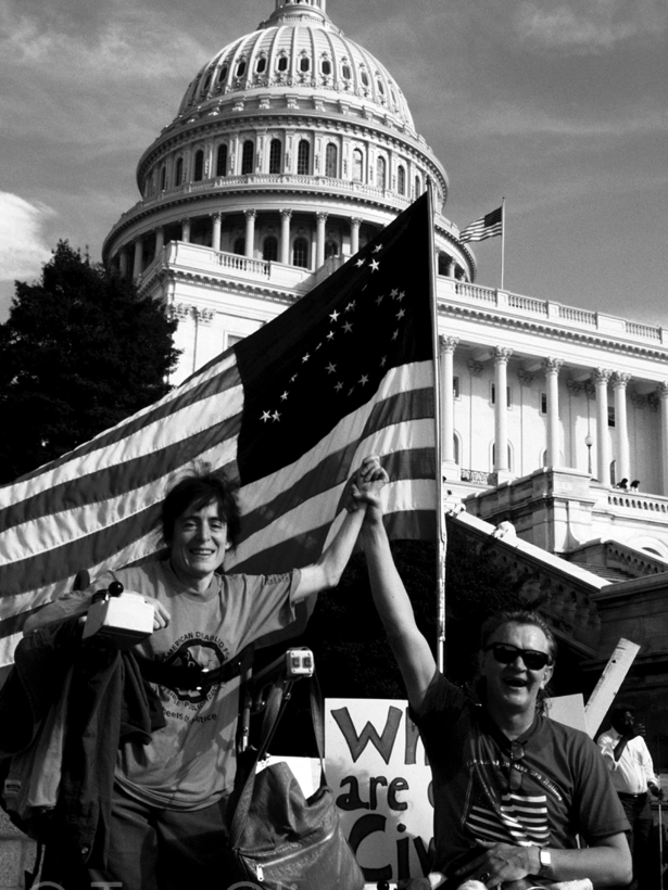 Two disability rights activists, one using a wheelchair and the other using a scooter, hold hands triumphantly in front of the U.S. Capitol. An American flag with a circle of stars waves behind them, symbolizing unity and progress.