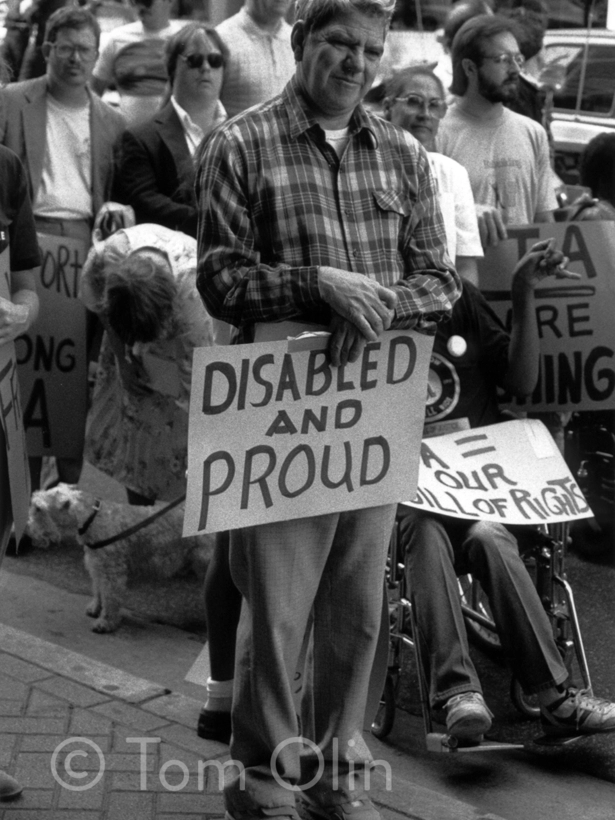 A man stands among a crowd during a disability rights protest, holding a sign that reads “Disabled and Proud.” People around him carry additional protest signs, and a person in a wheelchair is partially visible behind him.